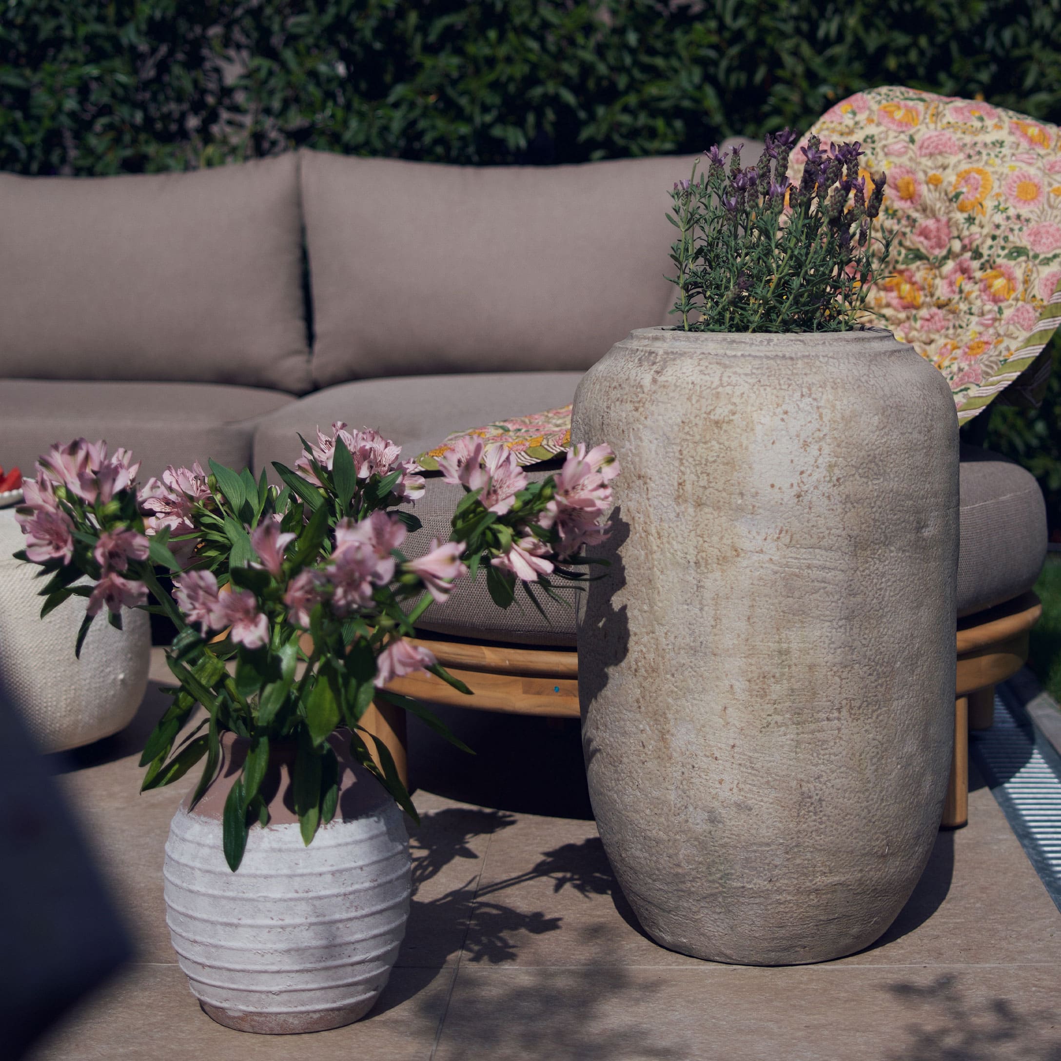 Grand et petit pot en pierre claire avec fleurs sur terrasse avec canapé gris et couverture colorée