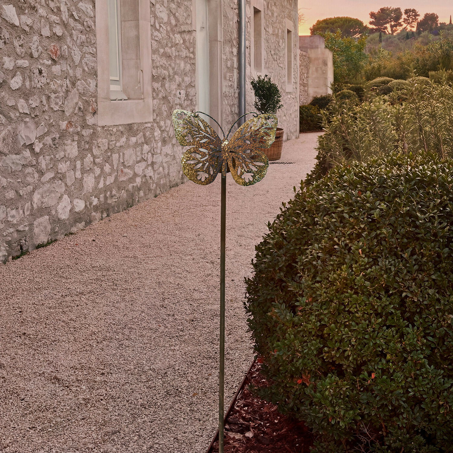 Décoration de jardin en métal en forme de papillon couleur laiton devant un mur en pierre et des buissons verts