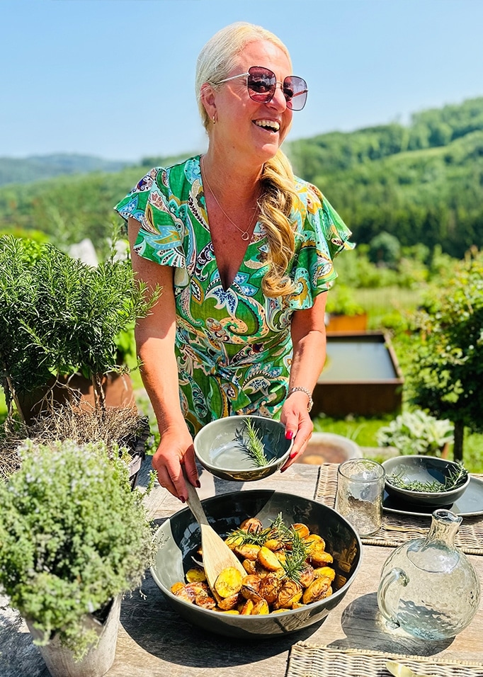 Femme en robe verte servant des pommes de terre rôties dans un bol en céramique en plein air avec des herbes et nature