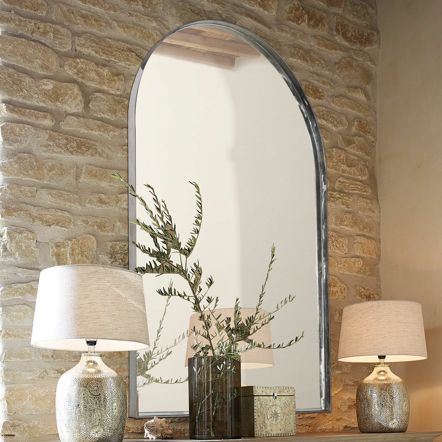 Miroir Kellin Grand miroir à cadre métallique devant un mur en pierre avec lampes et décorations sur une étagère en bois