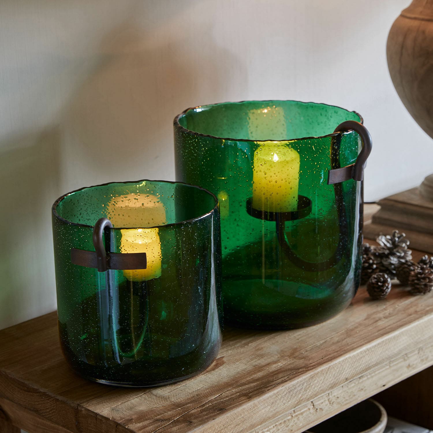 Lot de 2 photophores Ylva Ensemble de deux photophores en verre vert avec poignées métalliques sur une table en bois