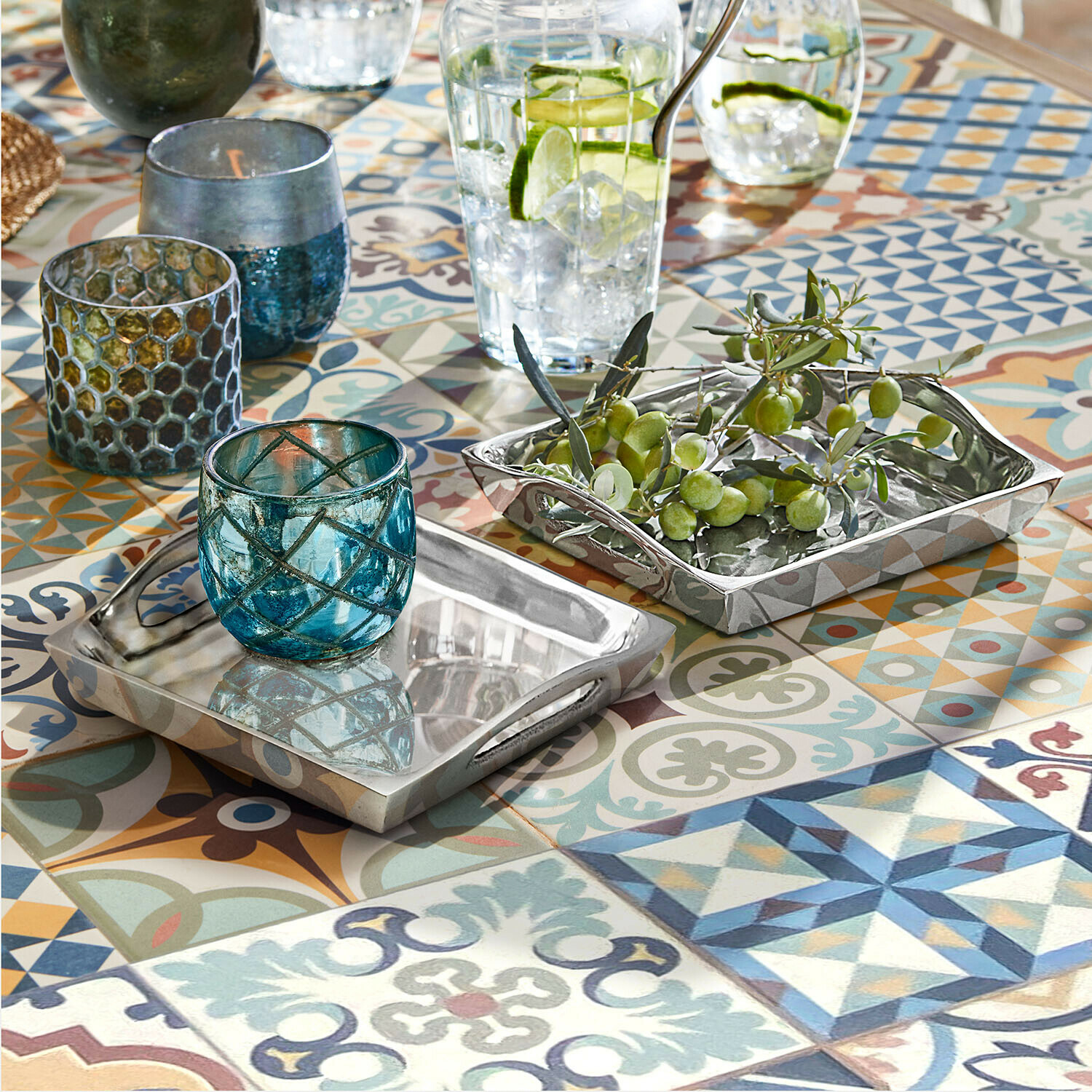 Plateaux métalliques argentés rectangulaires sur table colorée à motifs, bougeoirs en verre bleu et olives vertes