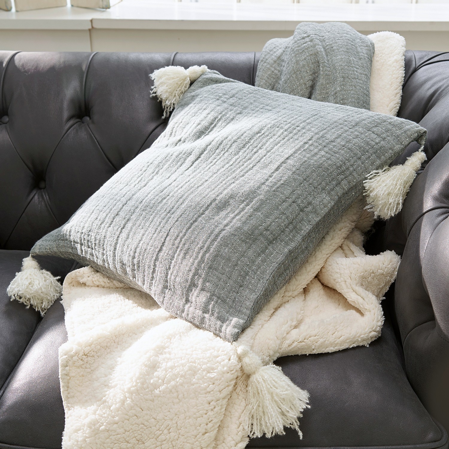 Coussin Malliévre Coussin gris à pompons et plaid crème doux sur canapé en cuir foncé pour décoration salon