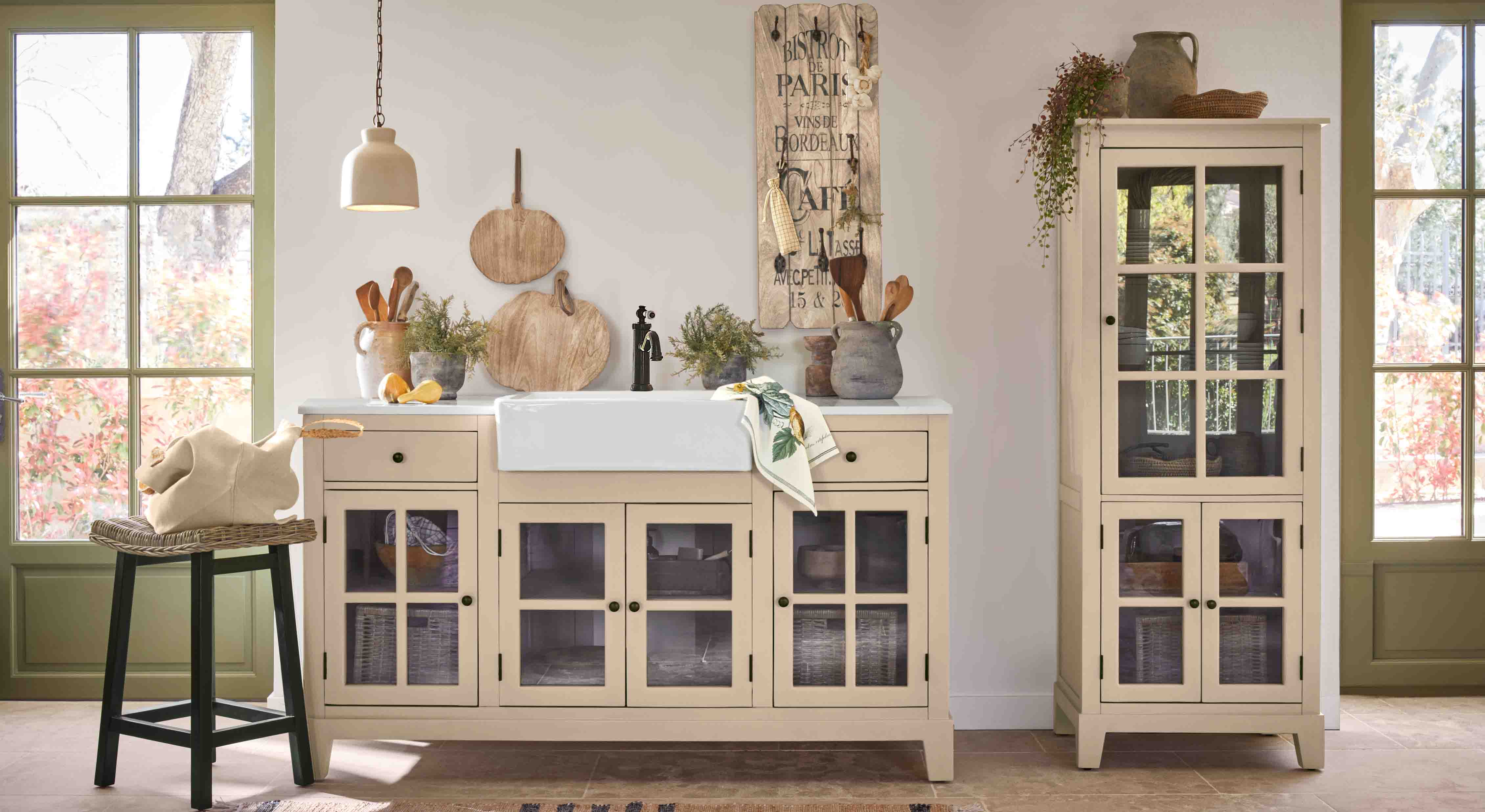 Cuisine rustique avec meubles beige à portes vitrées et évier en céramique blanc, décorations en bois et plantes