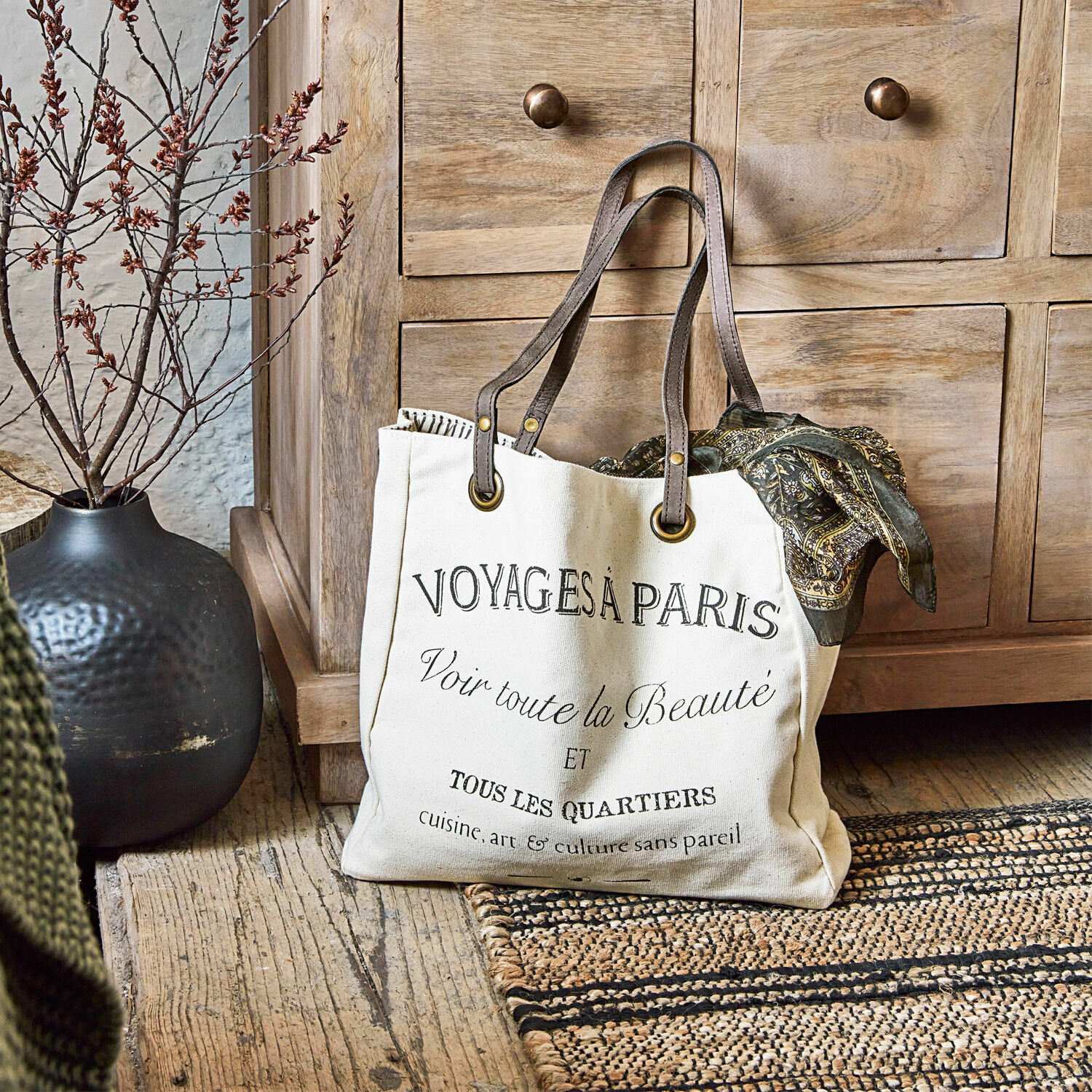 Cabas Voyage Sac en coton beige avec impression noire et anses marron posé sur un sol en bois rustique