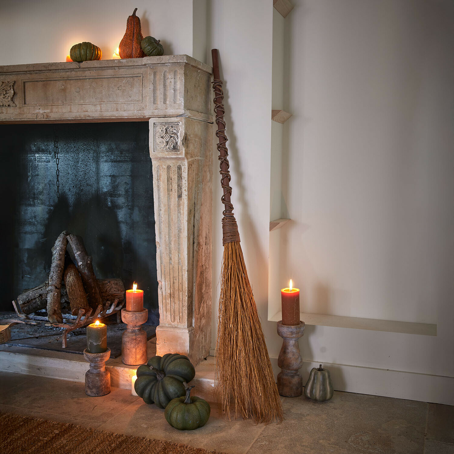 Balai décoratif Maerlinna Balai naturel en brindilles appuyé contre une cheminée en pierre avec citrouilles vertes et bougies allumées