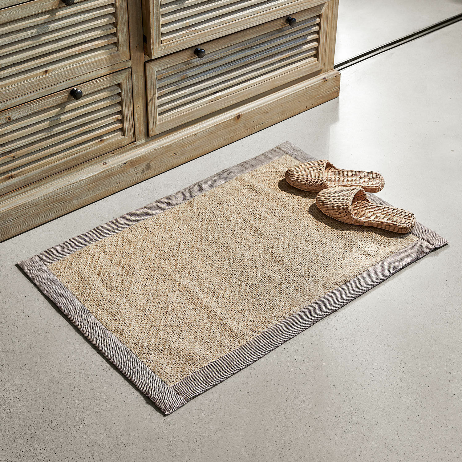 Tapis de bain Wilder Tapis beige en matériau naturel avec bordure grise sur sol clair près d'un meuble en bois