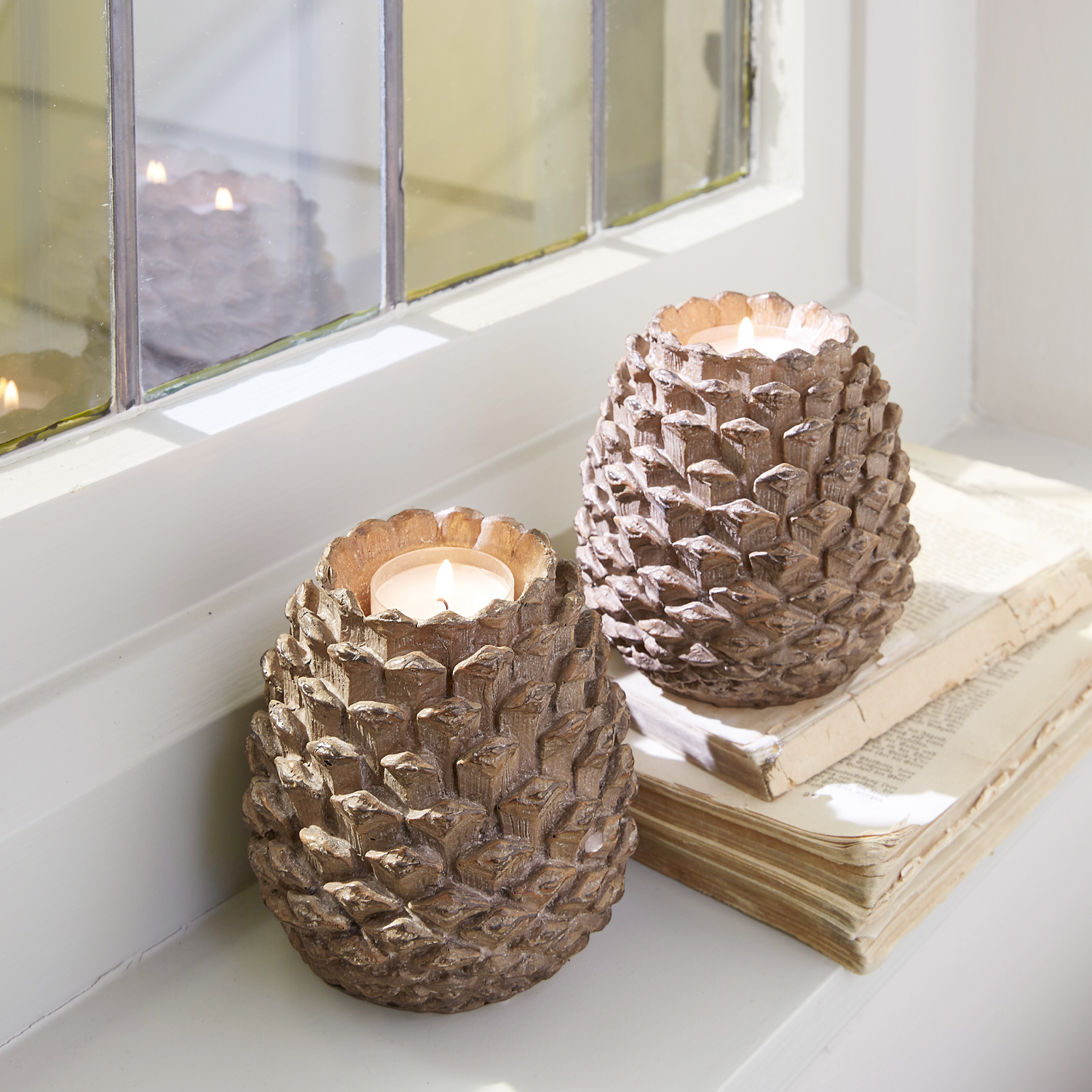 Deux bougeoirs en forme de pomme de pin en bois brun posés sur un rebord de fenêtre avec des livres