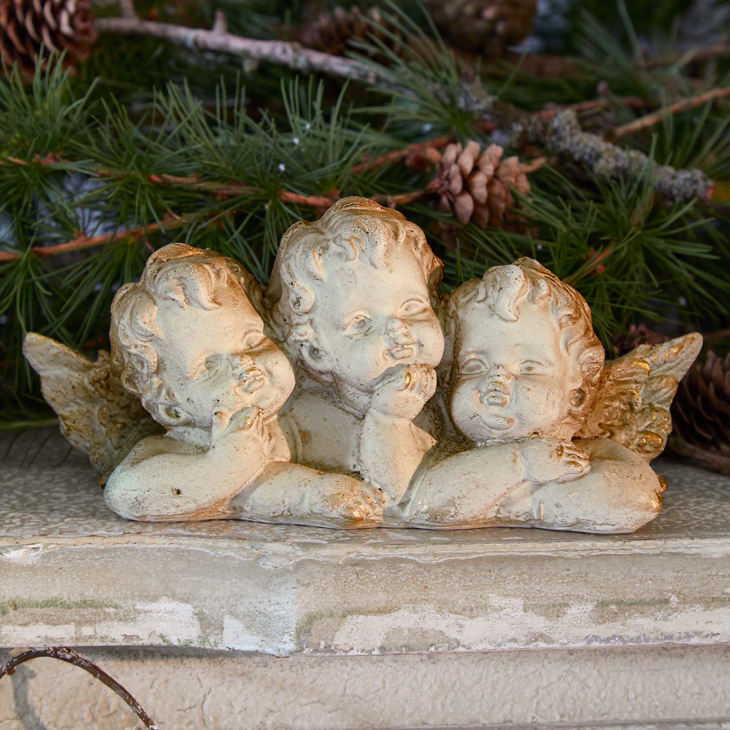 Figurine de trois petits anges en pierre claire sur fond de branches vertes et pommes de pin, décoration