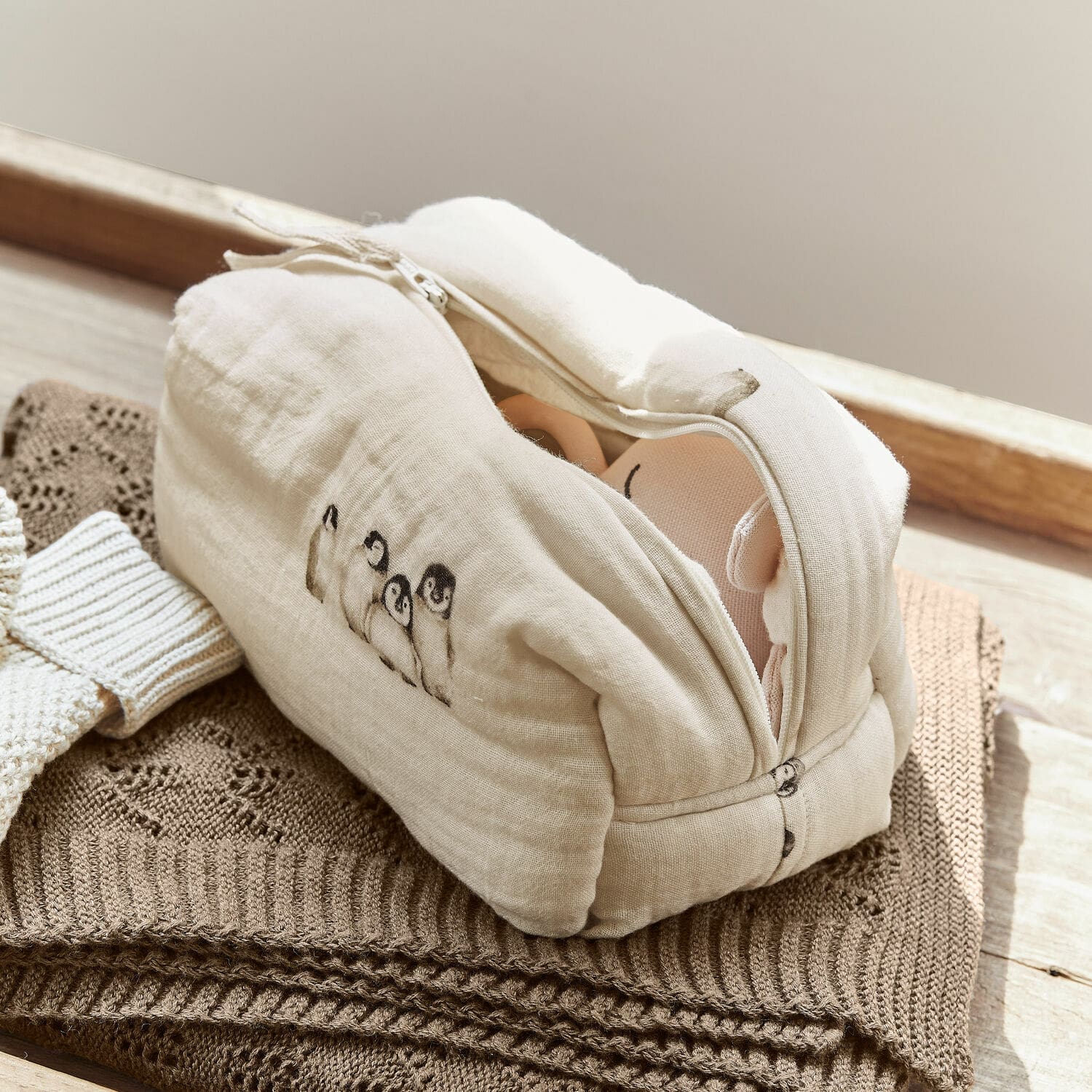 Pochette Jocienne Trousse de toilette beige en coton avec motif pingouins posée sur un tricot marron, rangement pratique