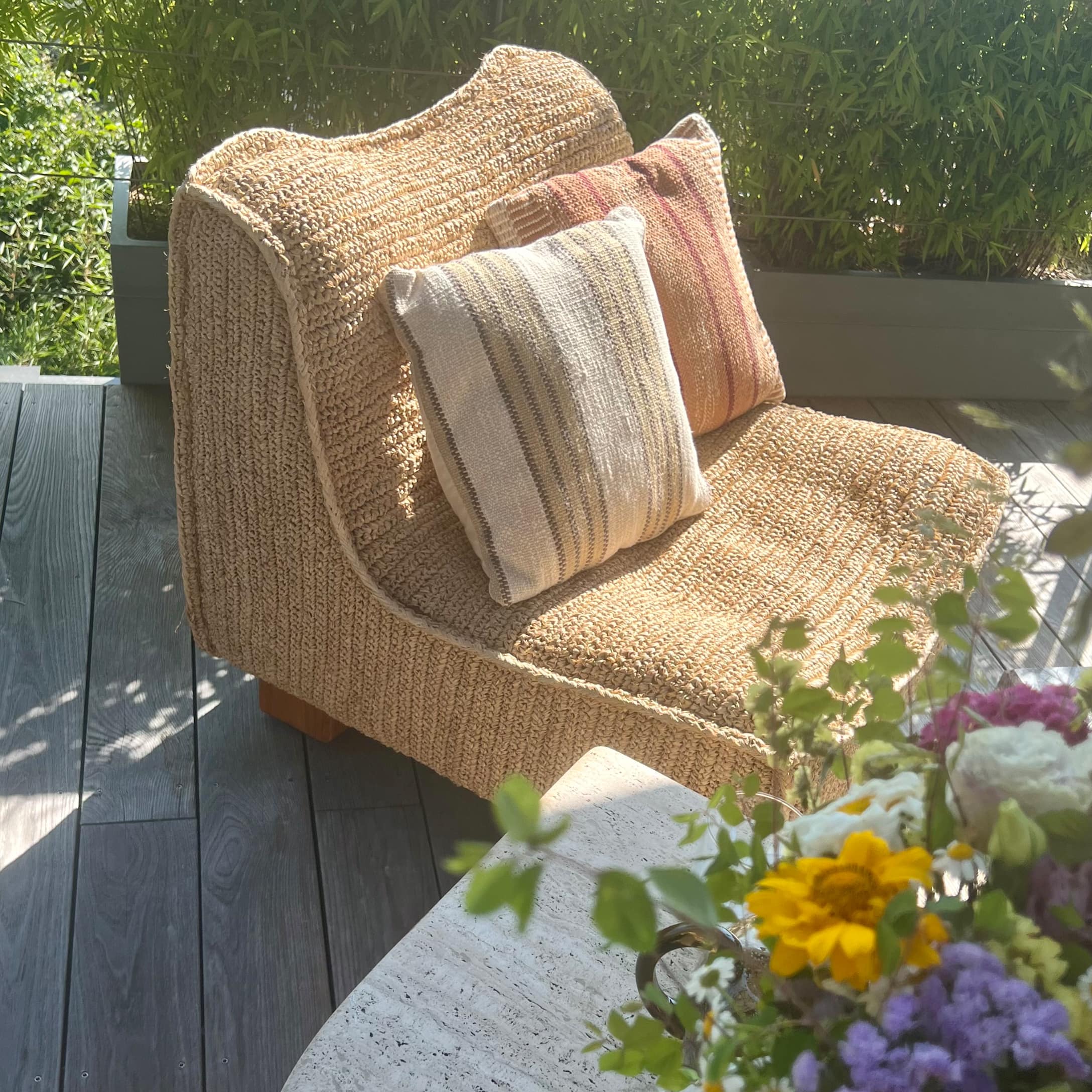 Fauteuil beige en matière tressée avec deux coussins décoratifs sur terrasse avec fleurs colorées