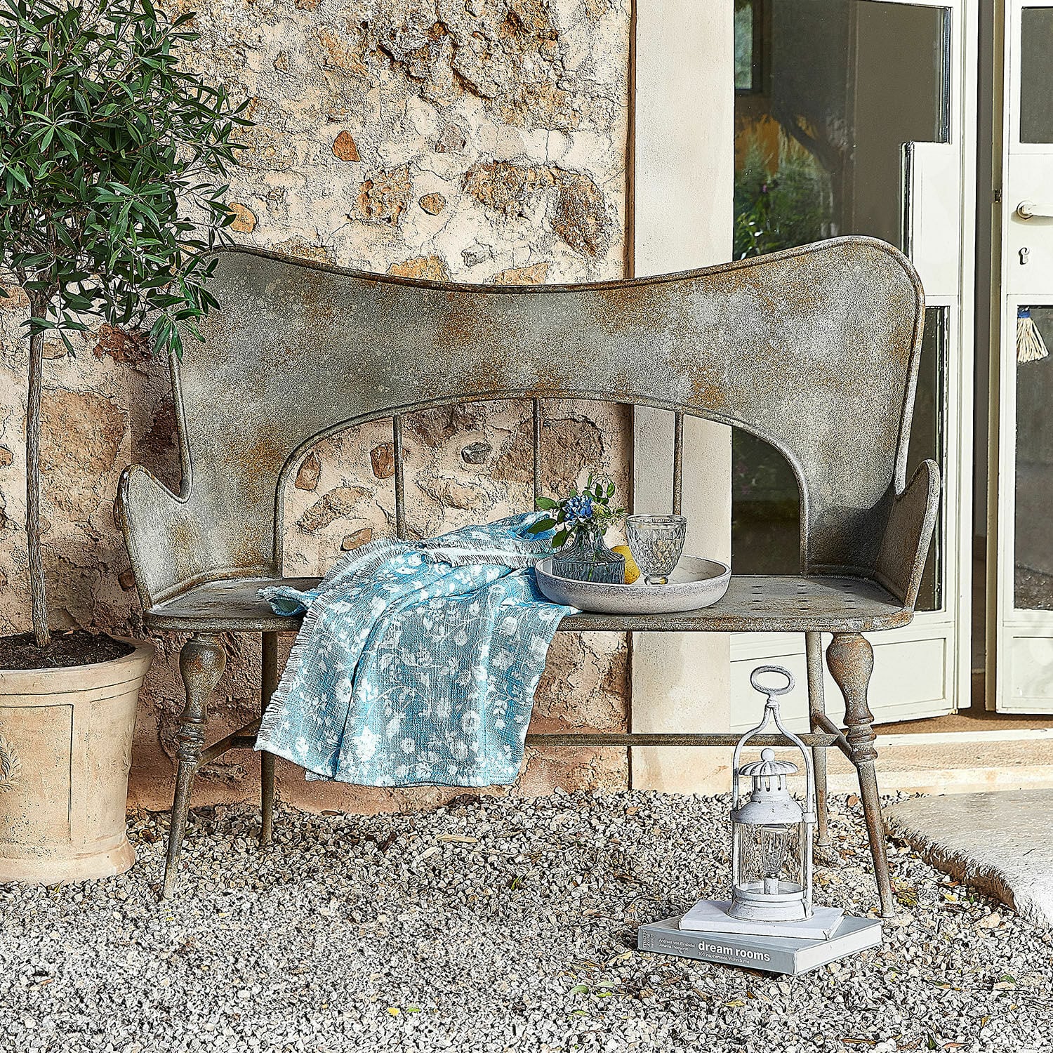 Banc Horivel Banc de jardin en métal patiné gris avec couverture bleue sur sol en gravier devant mur en pierre