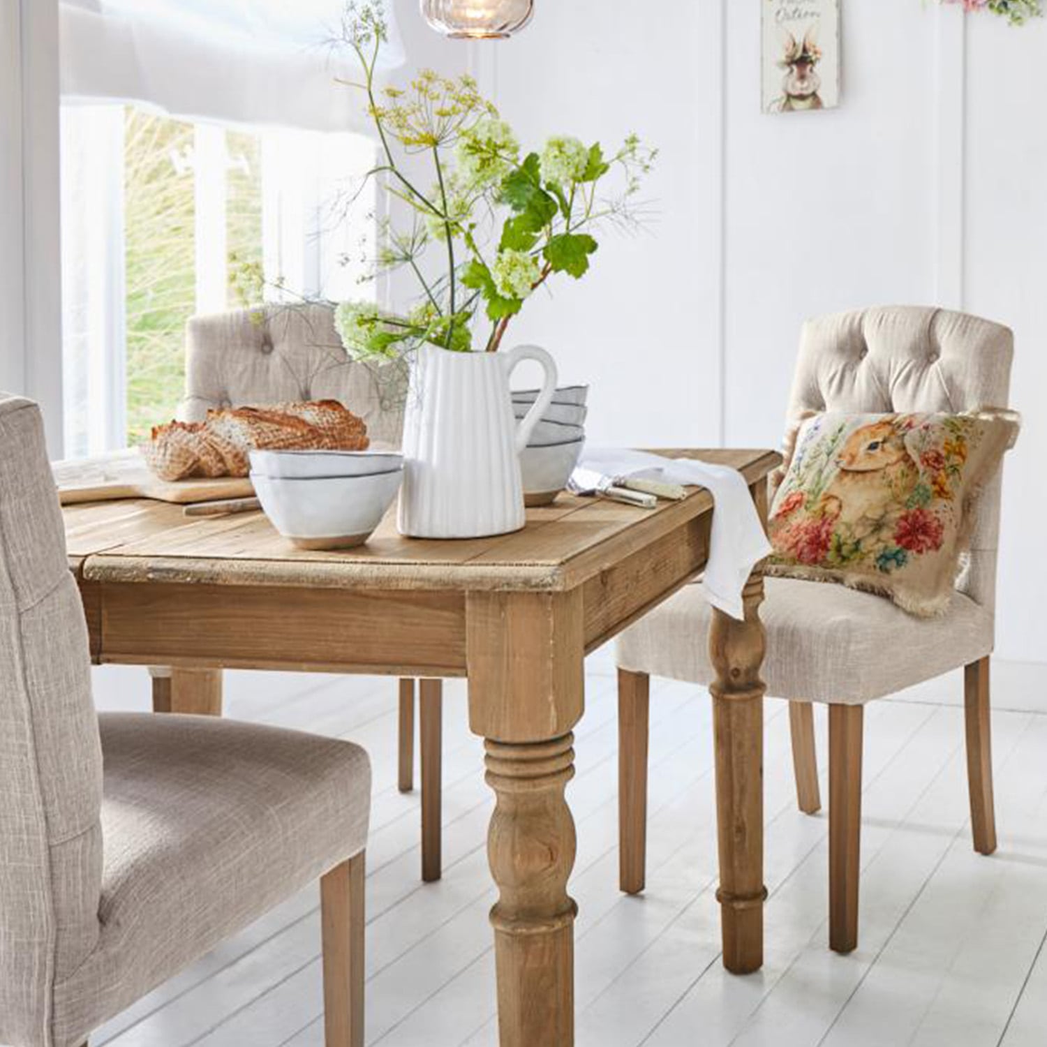 Table en bois clair avec pichet décoratif et bols, chaises rembourrées beige dans une salle à manger lumineuse