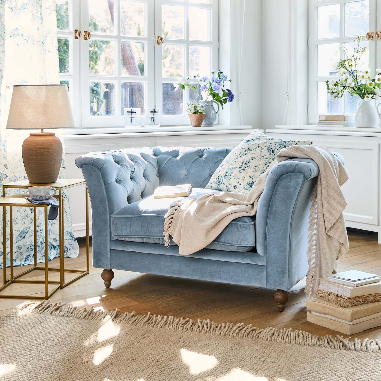 Canapé deux places en velours bleu avec coussin fleuri et plaid beige dans un salon lumineux au sol en bois