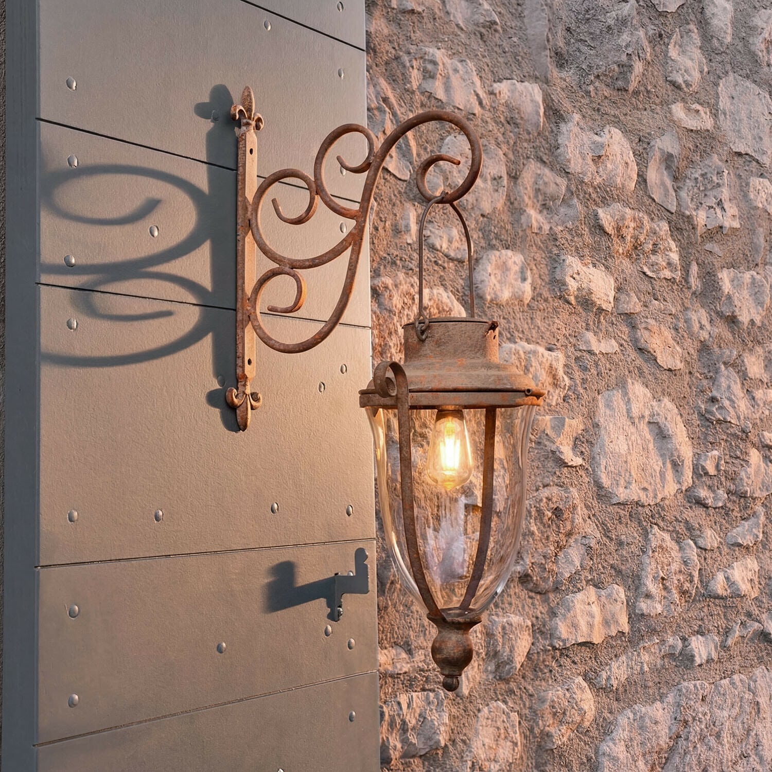 Lanterne solaire Volanaïs Applique murale extérieure en métal rouillé avec verre clair fixée sur mur en pierre, lumière allumée