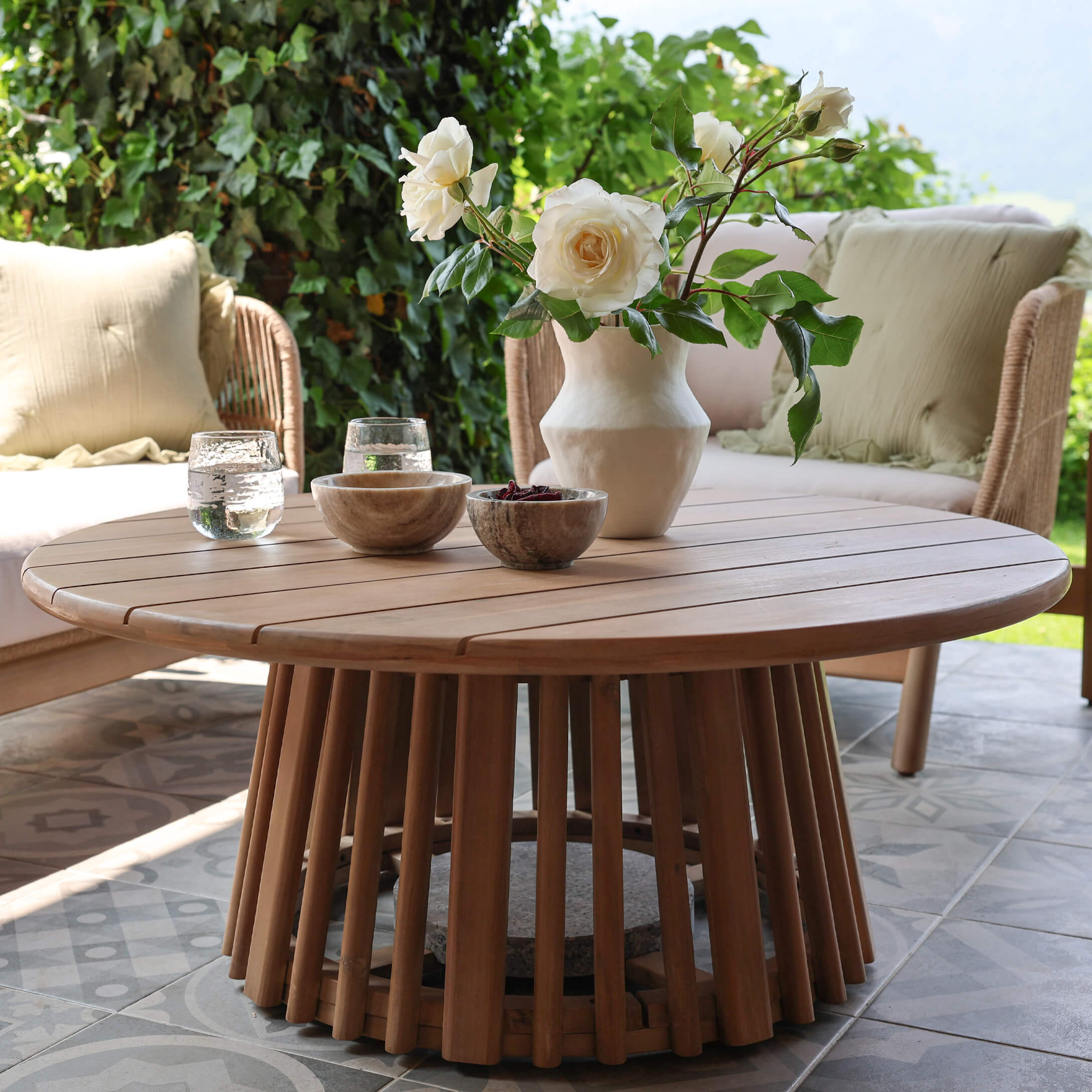 Table basse ronde en bois de teck naturel sur terrasse avec décoration florale et bols en pierre