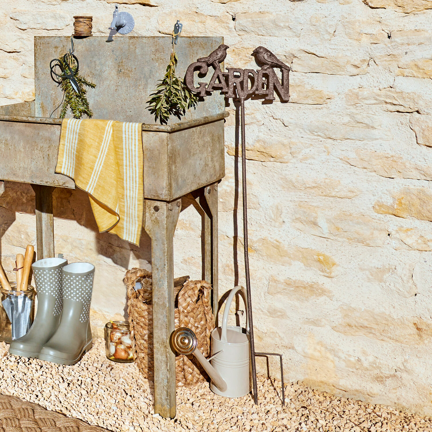 Pique décorative de jardin Girra Évier de jardin en métal avec torchon jaune et outils de jardinage devant un mur en pierre