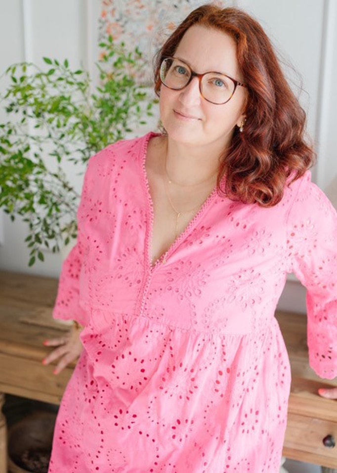 Femme en robe rose ajourée avec motif délicat, appuyée sur une table en bois avec plante en arrière-plan