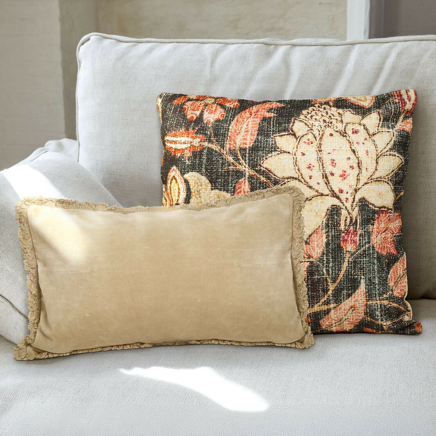 Lot de 2 housses de coussins Ambrayelle Deux coussins décoratifs sur un canapé gris clair, un beige avec franges, l'autre à motif floral coloré