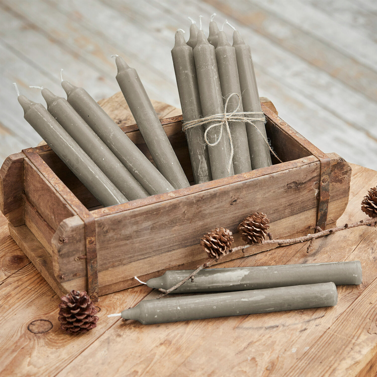 Lot de 12 bougies Bauceau Bougies de table grises en cire dans une caisse en bois sur une table rustique avec pommes de pin
