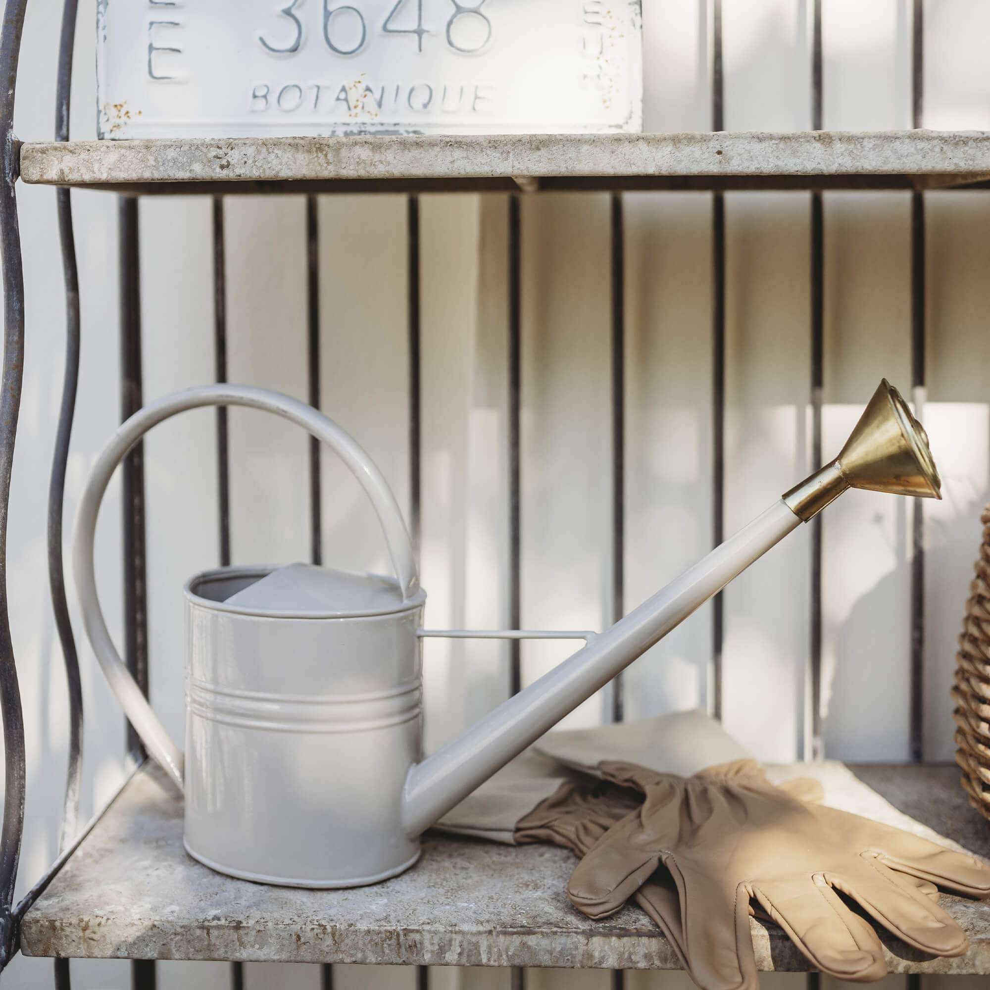 Arrosoir en métal beige avec bec en laiton et gants de jardinage posés sur une étagère en pierre
