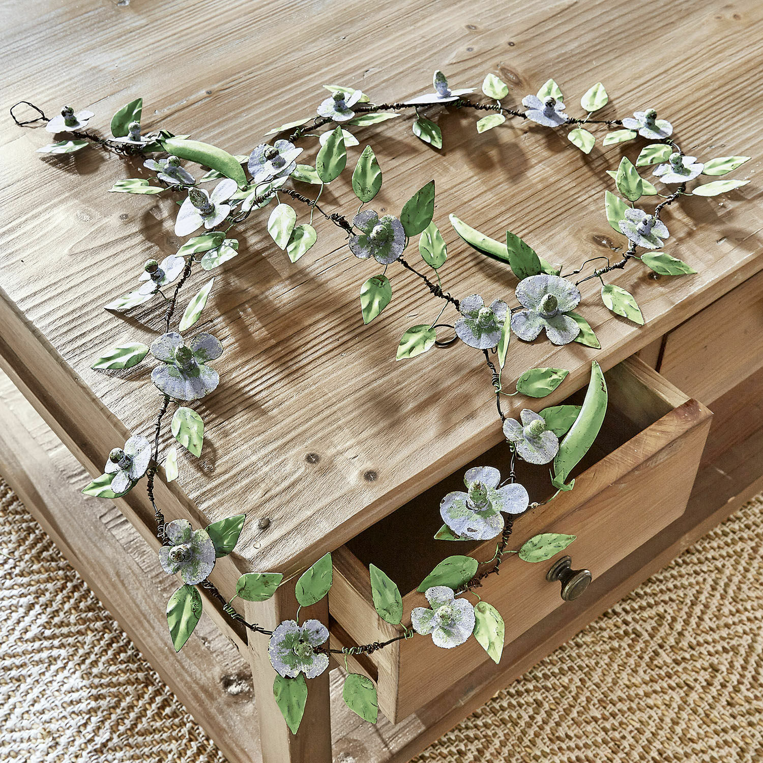Guirlande Cecilia Couronne décorative en métal avec fleurs et feuilles vertes et grises posée sur une table en bois clair
