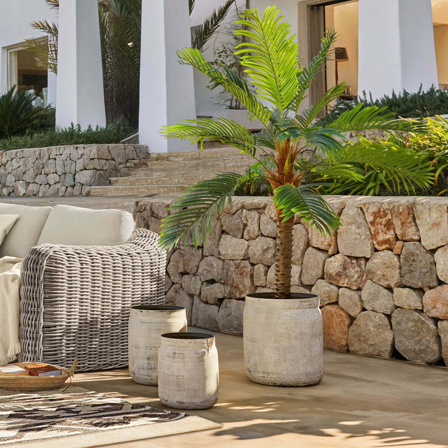 Ensemble de trois pots en béton clair sur terrasse avec palmier artificiel vert et mobilier en rotin