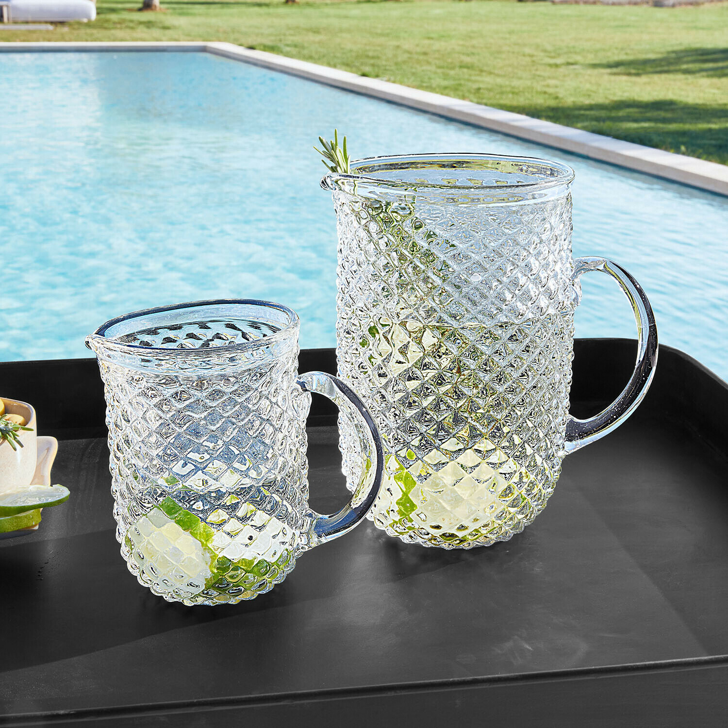 Carafe et pichet en verre transparent à motif diamant, garnis de tranches de citron vert, posés sur plateau noir près de la piscine