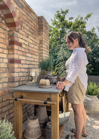 Femme en chemise blanche et short beige près d'une table de jardin en bois avec plateau en béton devant un mur en briques et des plantes