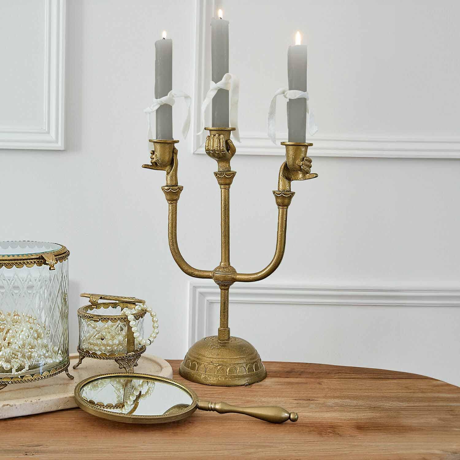 Chandelier doré à trois branches avec bougies grises tenues par des mains, décor de table avec miroir et boîtes