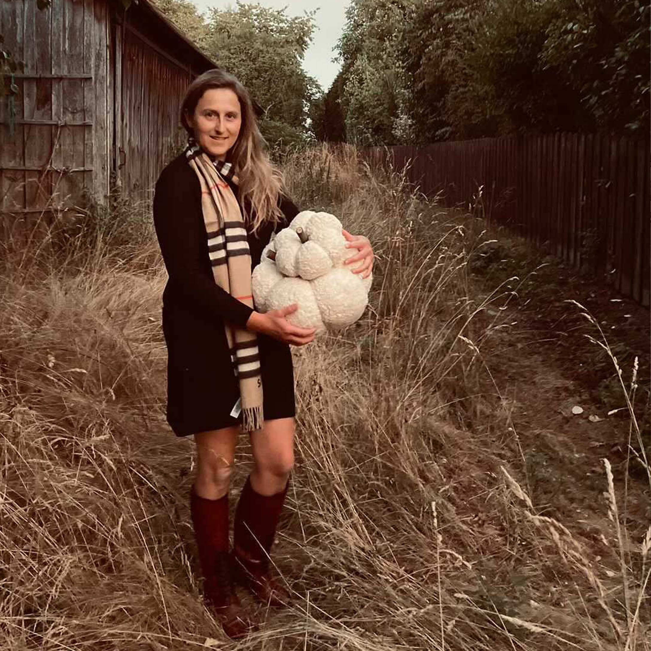 Femme en robe noire et écharpe tenant une citrouille décorative blanche dans un champ d'automne