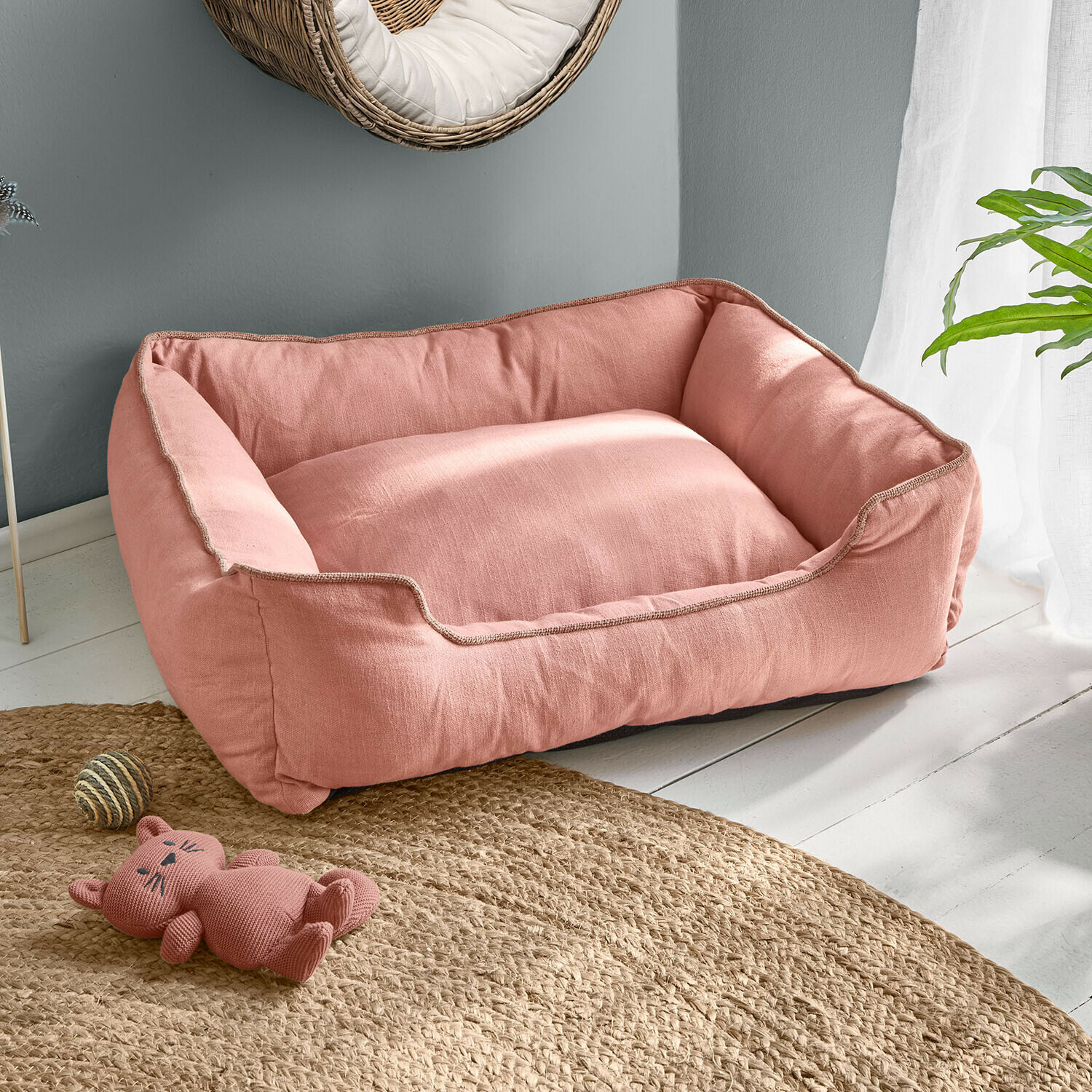 Panier pour chien rose en tissu doux sur sol clair avec jouet chat et tapis en jute
