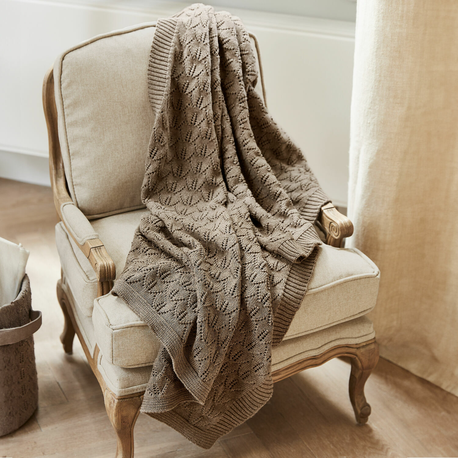 Couverture pour bébé Ernival Plaid beige en coton tricoté posé sur un fauteuil crème en bois clair dans un intérieur lumineux
