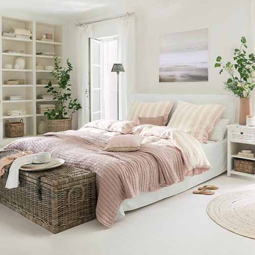 Chambre chaleureuse avec lit en linge blanc et couverture rose, décorations naturelles et coffre en osier