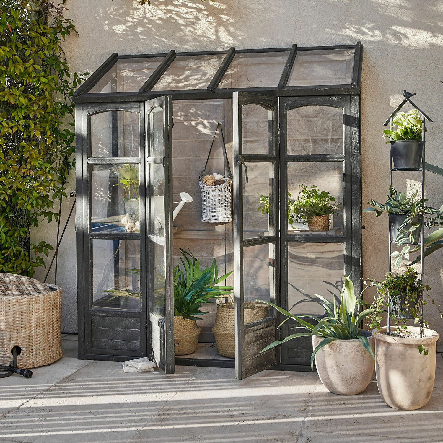 Serre Abilenne Petite serre de jardin avec cadre métallique noir et plantes en pots sur une terrasse ensoleillée