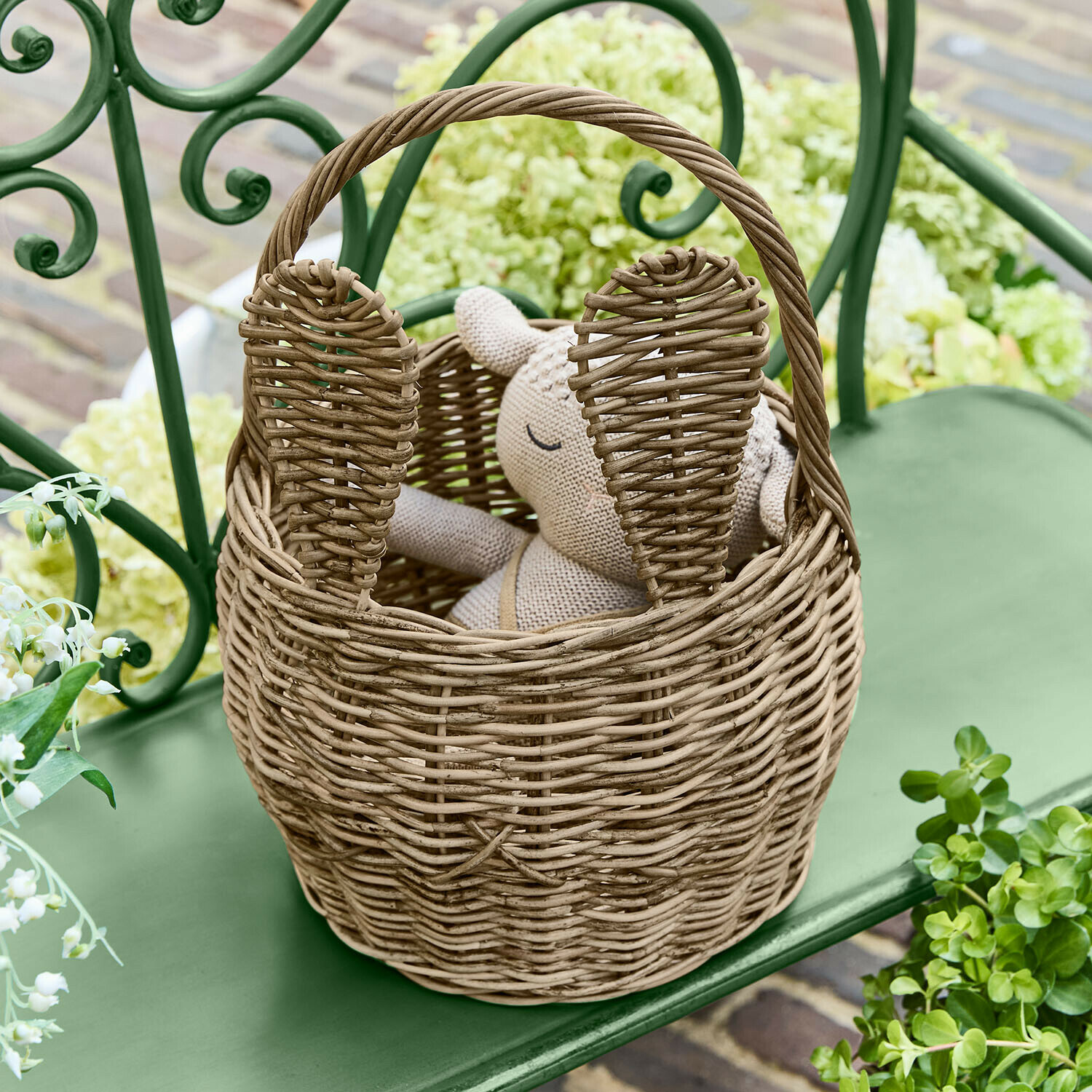 Panier Whimsywood Panier en osier naturel en forme de lapin avec oreilles, posé sur un banc vert dans un jardin