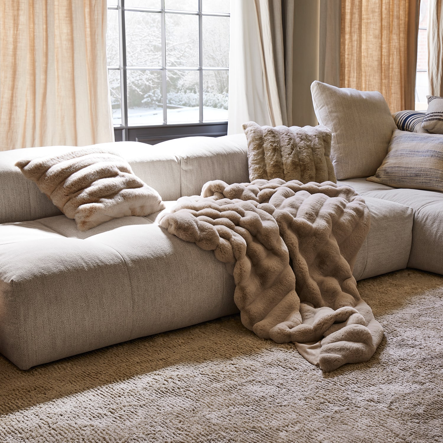 Plaid et coussins en fausse fourrure beige sur canapé clair dans un salon chaleureux avec vue sur jardin enneigé