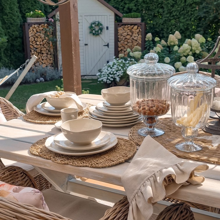 Service de vaisselle en céramique blanche sur table en bois clair avec bocaux en verre décoratifs dans un jardin