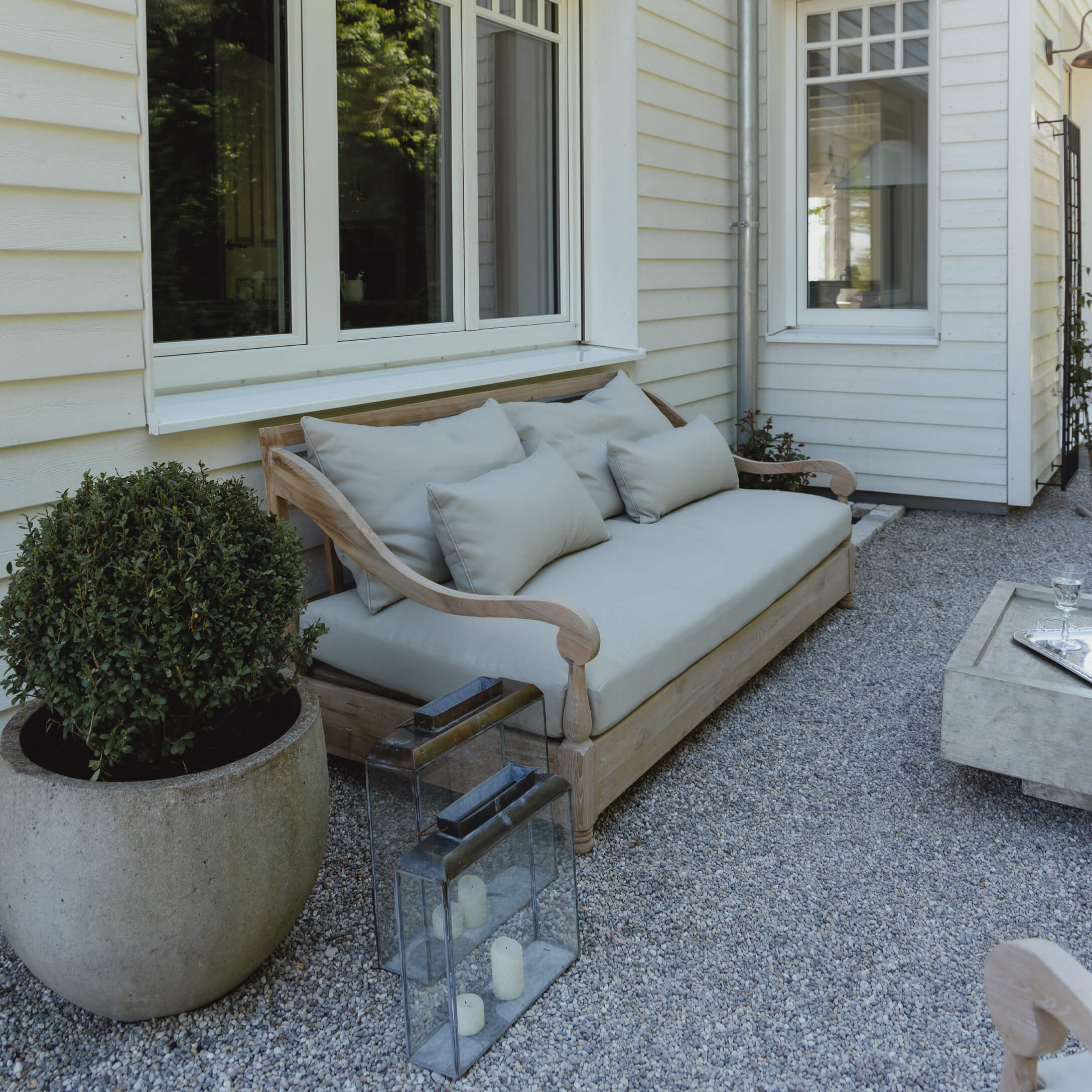Canapé d'extérieur en bois avec coussins gris clair sur patio en gravier devant maison blanche