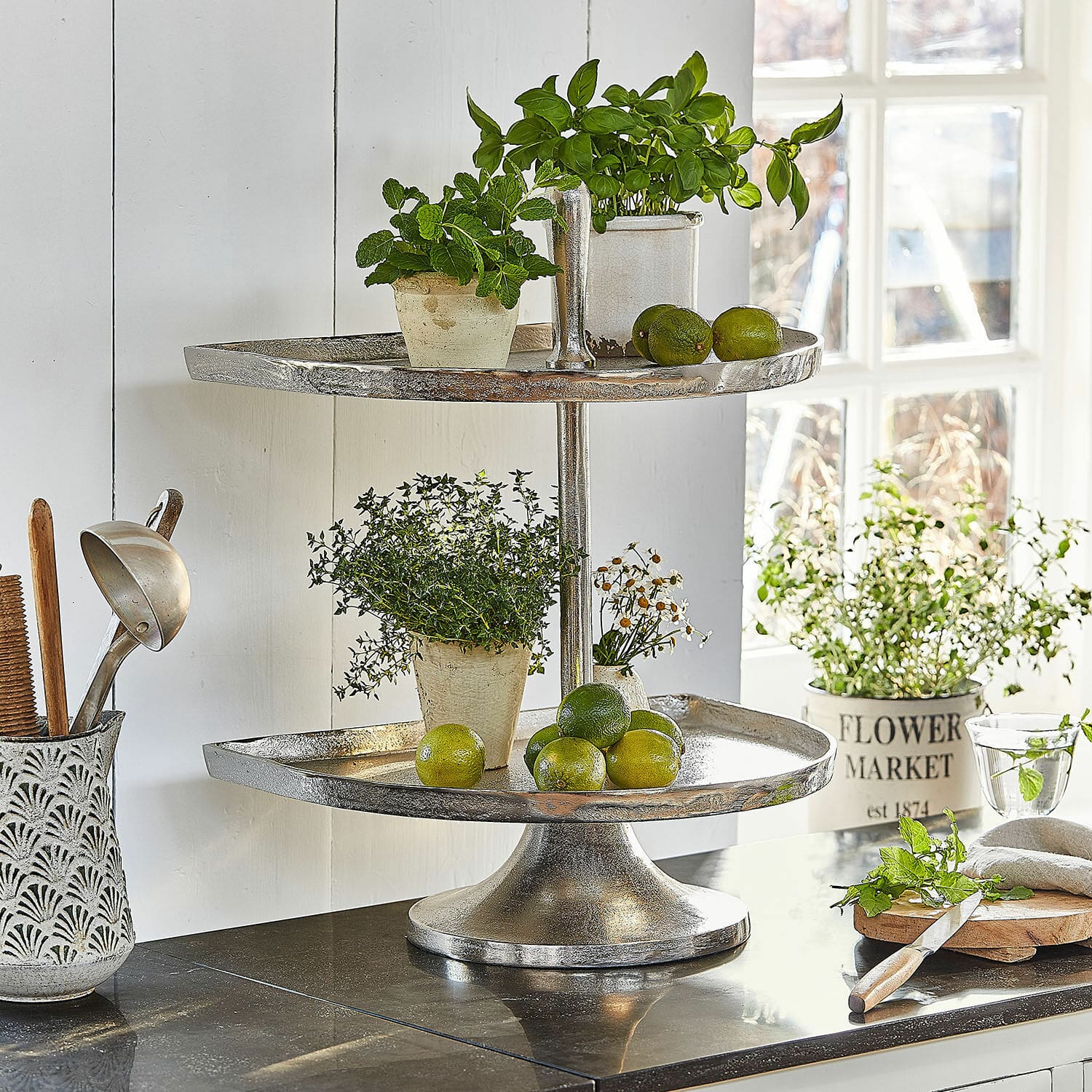 Présentoir à étages Quiralen Présentoir à deux niveaux en métal argenté avec herbes fraîches et citrons verts sur un plan de travail de cuisine