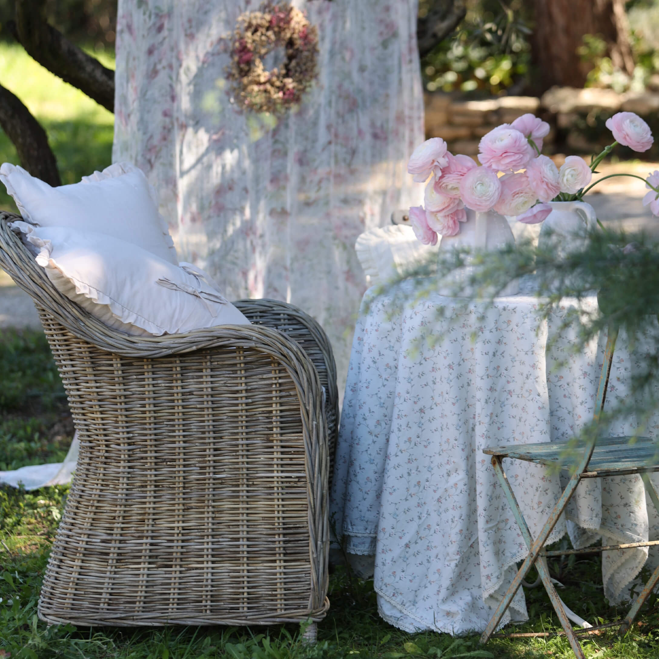 Fauteuil en osier avec coussins blancs à côté d'une table avec nappe fleurie et fleurs roses dans un jardin