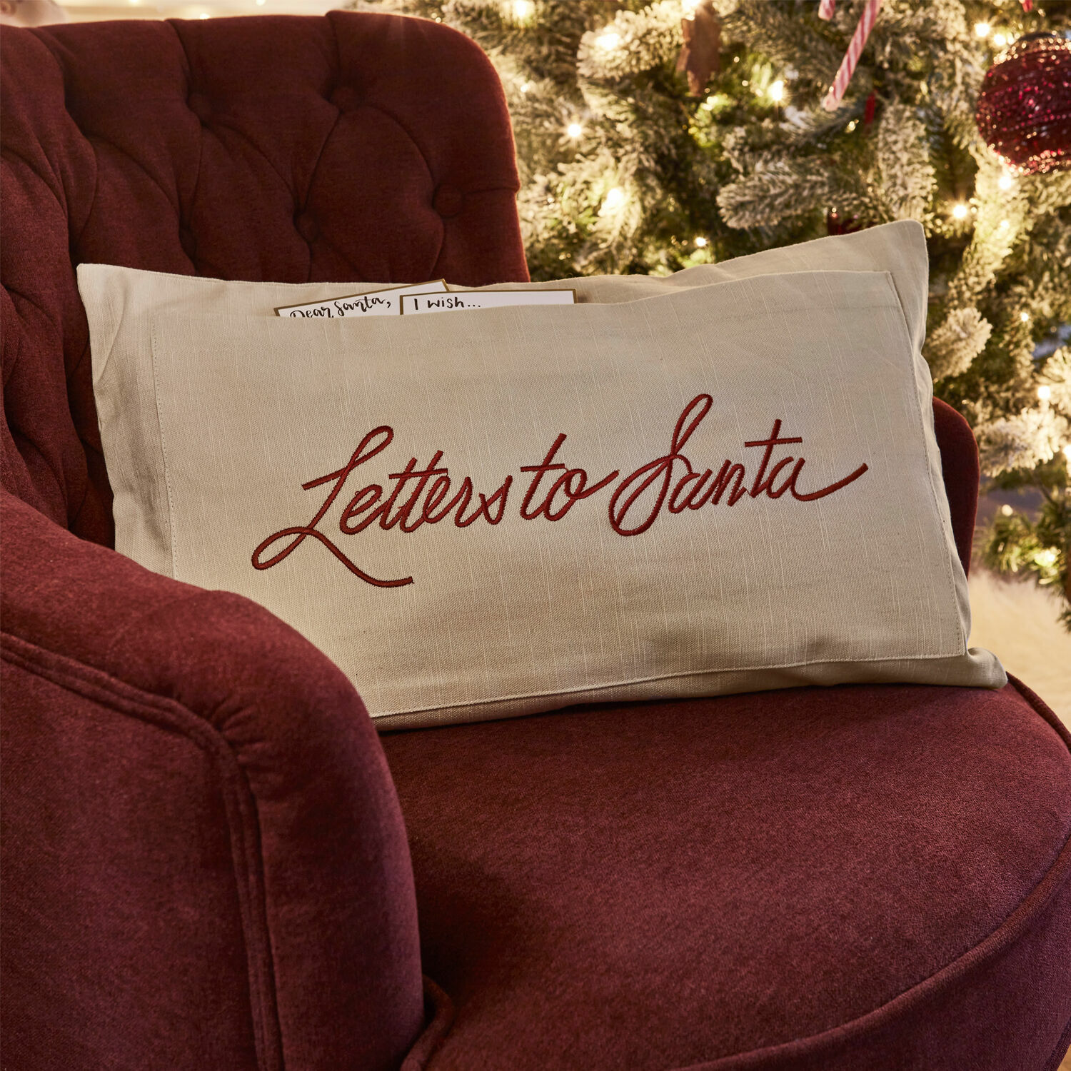 Housse de coussin Welenzas Coussin beige en lin avec inscription rouge Letters to Santa sur fauteuil bordeaux près d'un sapin de Noël