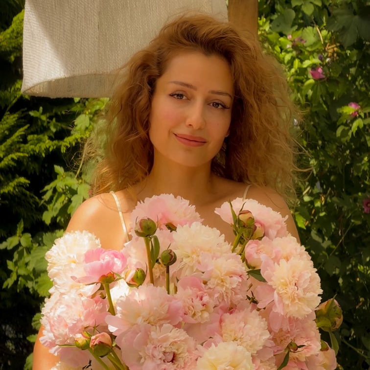 Femme aux cheveux longs tenant un grand bouquet de pivoines roses dans un jardin verdoyant