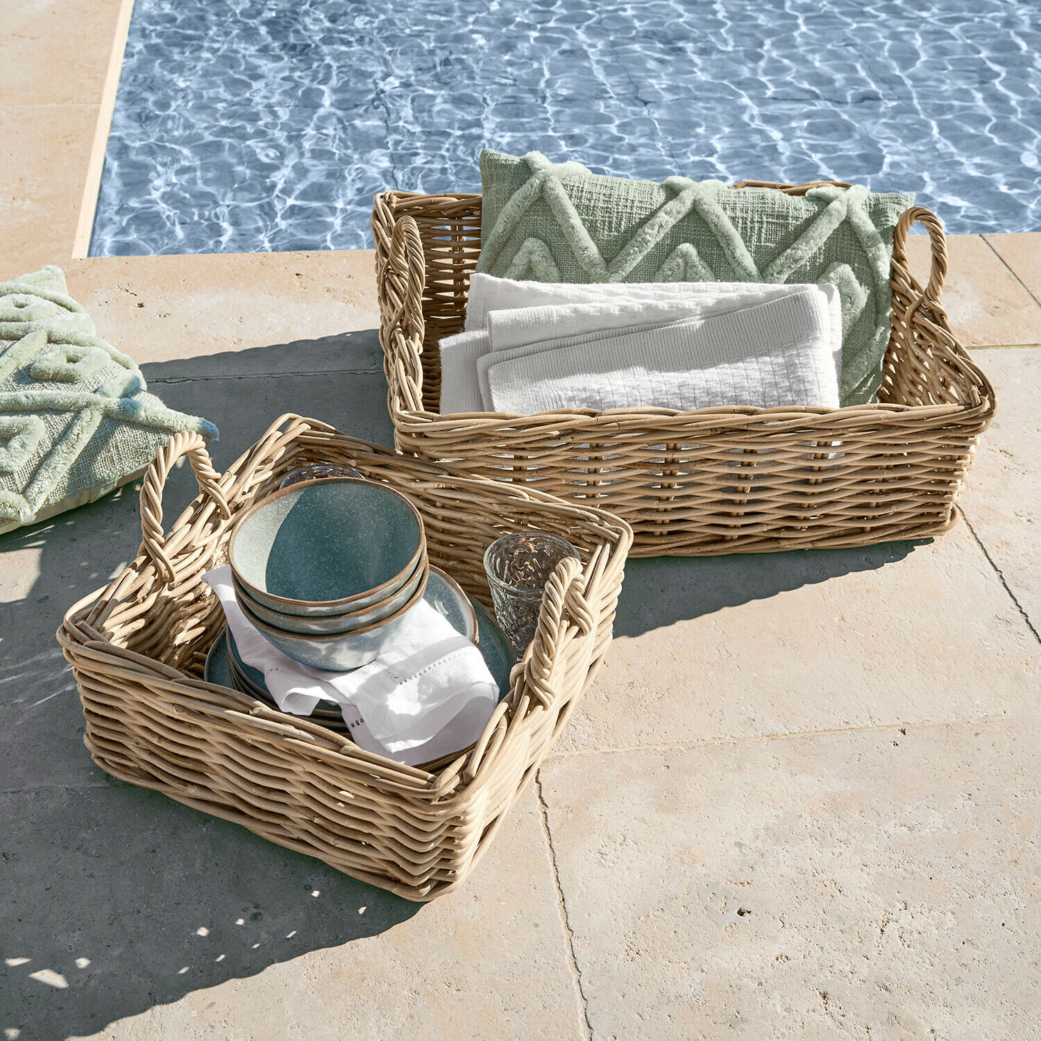 Deux paniers en osier avec poignées, garnis de coussins verts et serviettes blanches au bord de la piscine