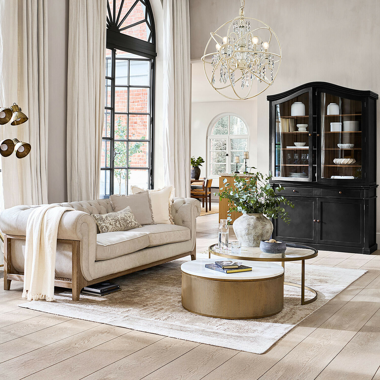 Salon chaleureux avec canapé beige, table basse dorée et vitrine noire sur fond de rideaux clairs et parquet en bois