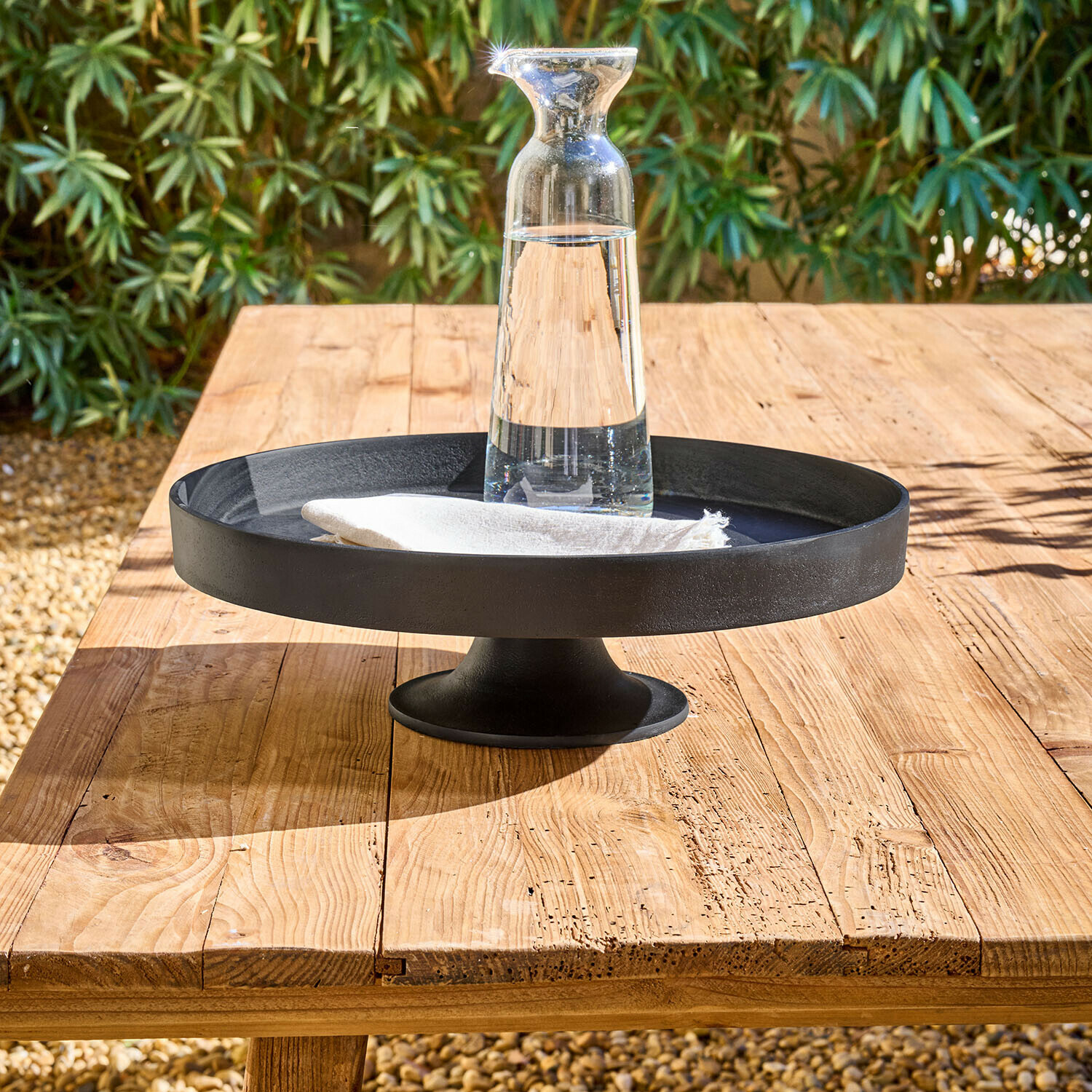 Présentoir Varelle Plateau noir en métal sur pied avec carafe en verre transparent et serviette blanche sur table en bois