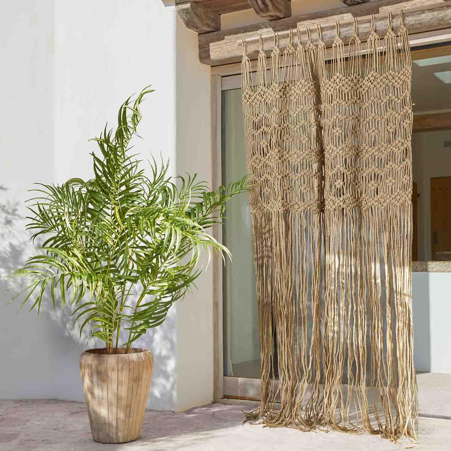 Rideau macramé beige en coton avec longues franges devant une porte-fenêtre, plante en pot à côté