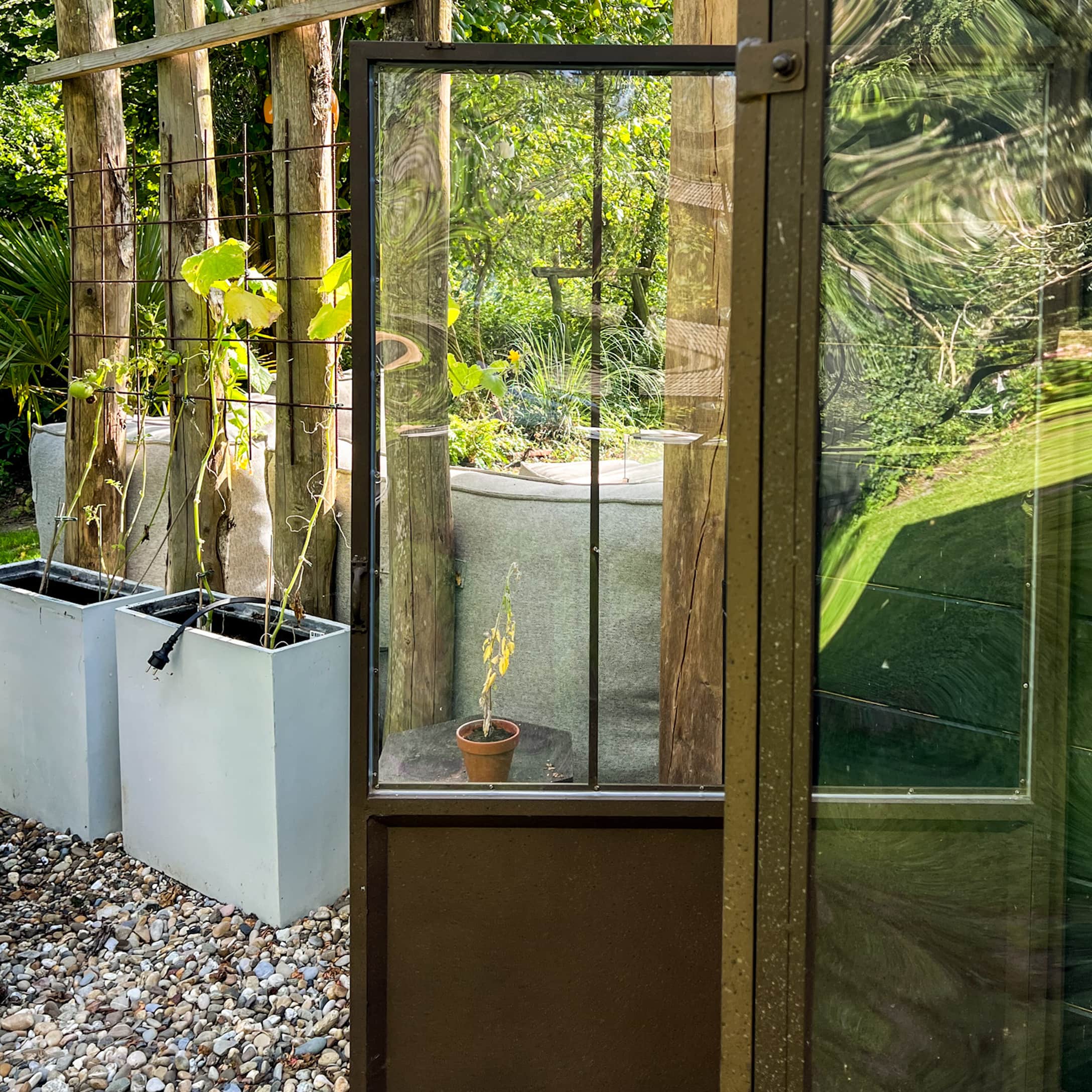 Porte de jardin en verre avec cadre métallique entourée de plantes en pots sur un sol en gravier