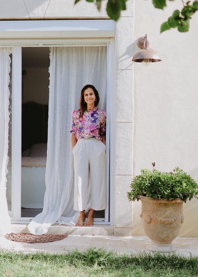 Femme en blouse fleurie et pantalon blanc debout dans une porte avec un rideau blanc et un pot de plante verte