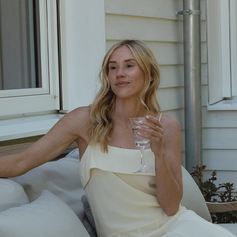 Femme en robe crème assise dehors, se détendant avec un verre à la main sur un canapé de jardin